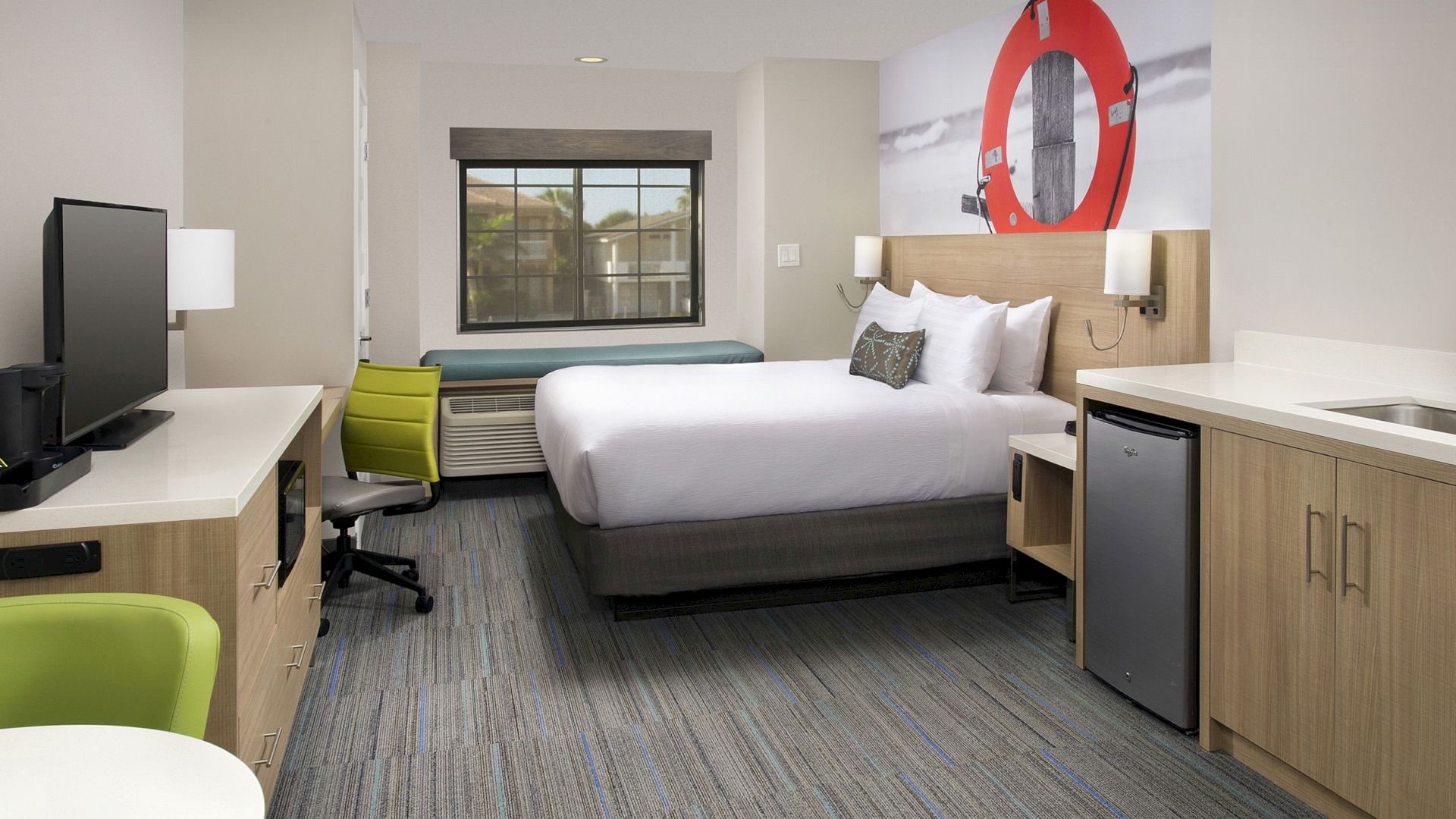 A modern hotel room features a bed, TV, small fridge, desk, and window seat with a decor of a life preserver on the wall.