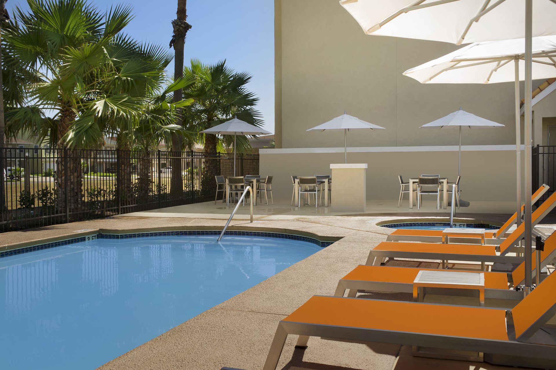 A poolside area with orange lounge chairs, white umbrellas, palm trees, and a seating area near a beige building is shown.