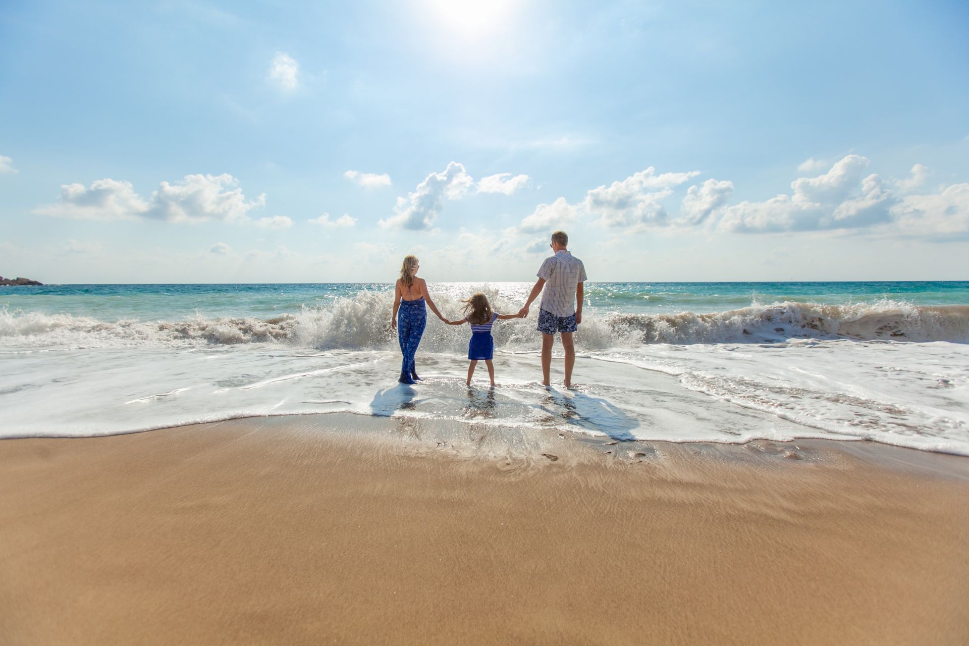 Three people, two adults and a child, are holding hands facing the ocean on a sunny beach with waves approaching.
