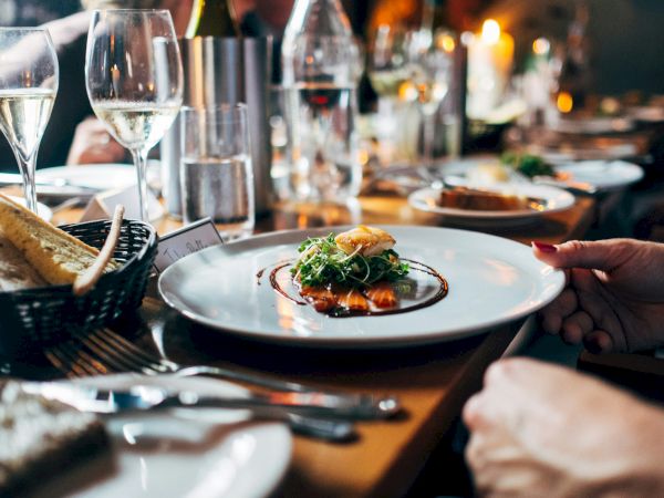 The image shows an elegant dining setup with gourmet dishes, wine glasses, and bread on a table in a restaurant setting.