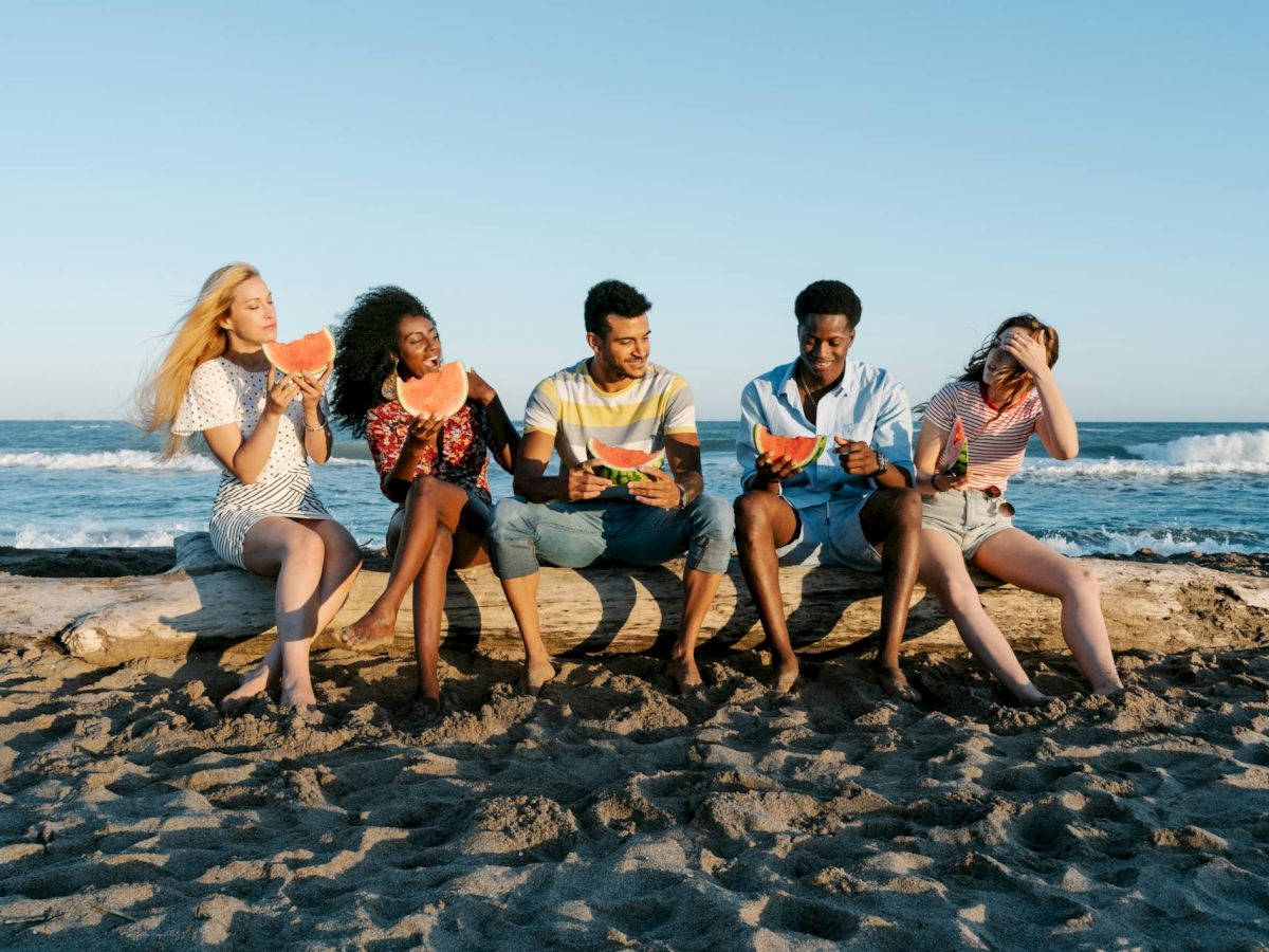 A diverse group of five friends sits on a sandy beach by the ocean, enjoying snacks and drinks under a clear blue sky.