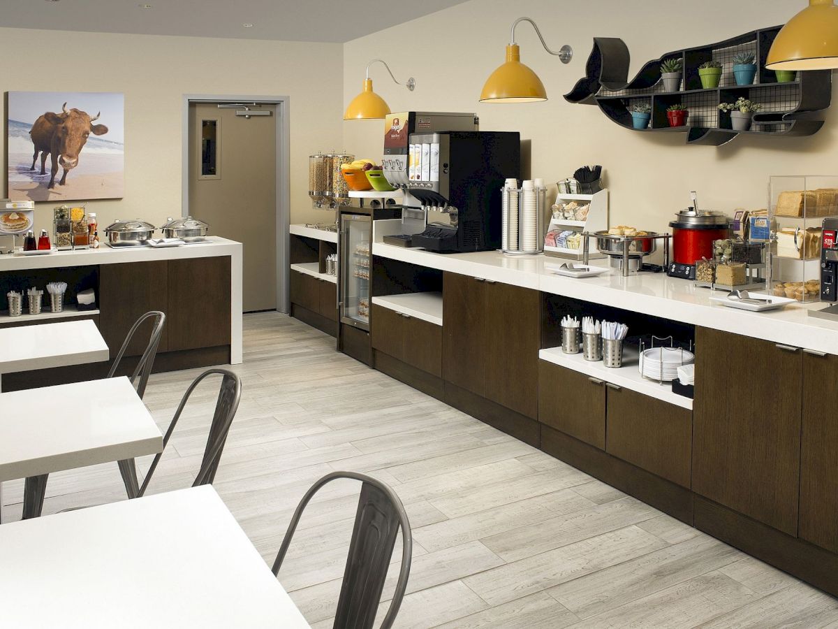 A modern caf&eacute;-style kitchen with white countertops, dark wood cabinets, industrial chairs, hanging yellow lights, and a coffee station with appliances and shelves.
