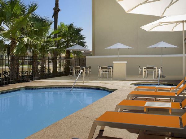 A sunny poolside area with blue water, orange lounge chairs, white umbrellas, and a beige wall behind.