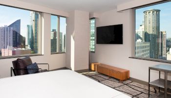 A modern hotel room with large windows offering a city view, a bed, a chair, a TV mounted on the wall, and a small table.