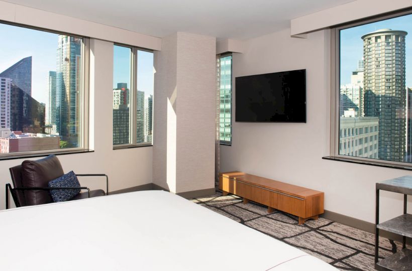 A modern hotel room with large windows offering a city view, a bed, a chair, a TV mounted on the wall, and a small table.