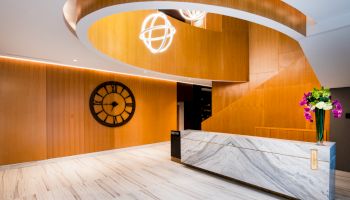 A modern lobby with a marble desk, wooden accents, geometric lights, a large clock, and a vase of flowers.