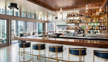 The image shows a modern bar with a wooden counter, barstools, and shelves stocked with various liquor bottles, in a high-rise setting.