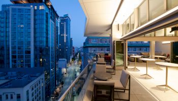A modern urban balcony with stylish seating overlooks a cityscape of tall buildings and illuminated streets at dusk.