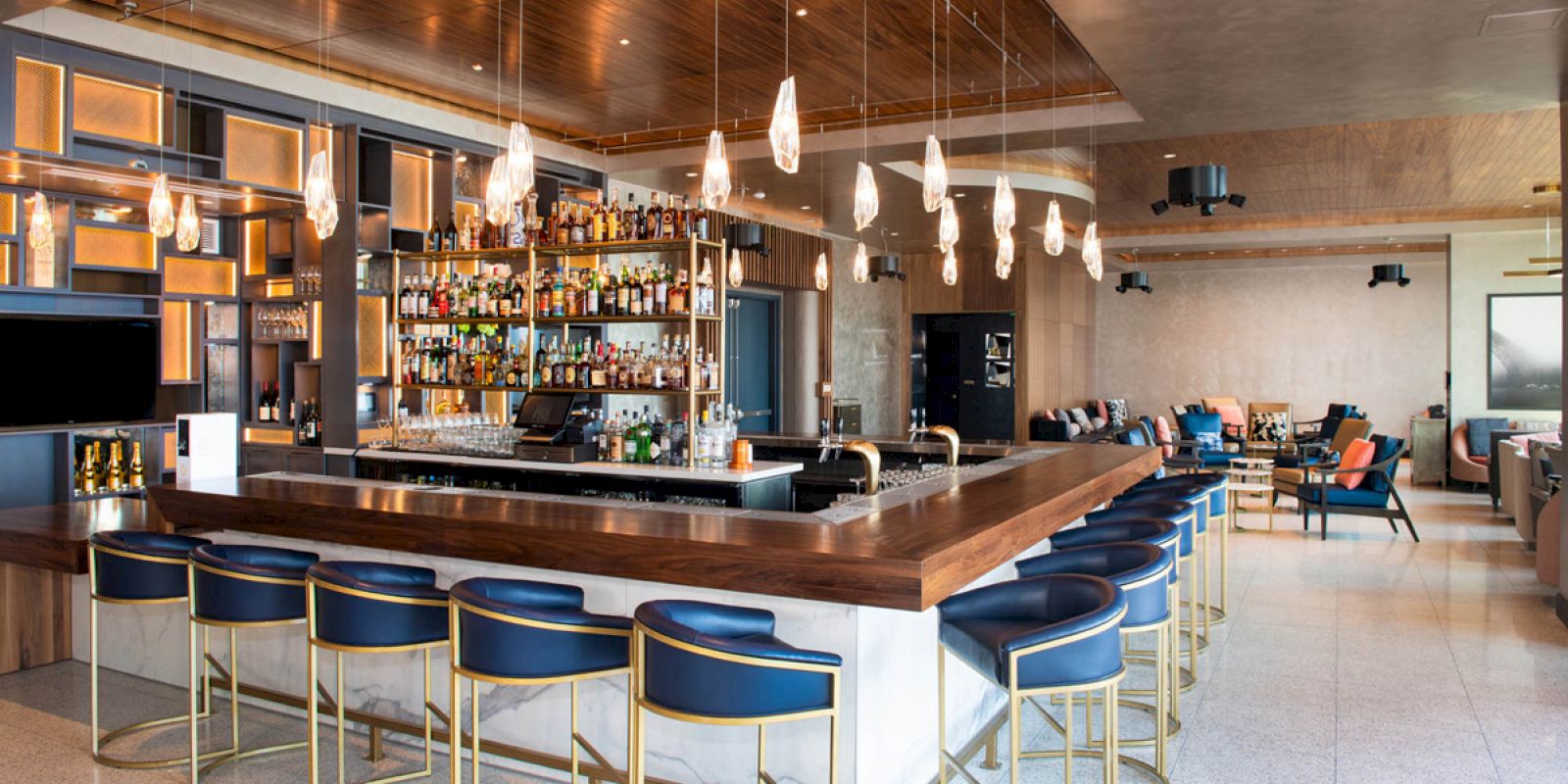 The image shows a modern bar with a central counter, stylish bar stools, hanging lights, and a stocked shelf, surrounded by cozy seating areas.