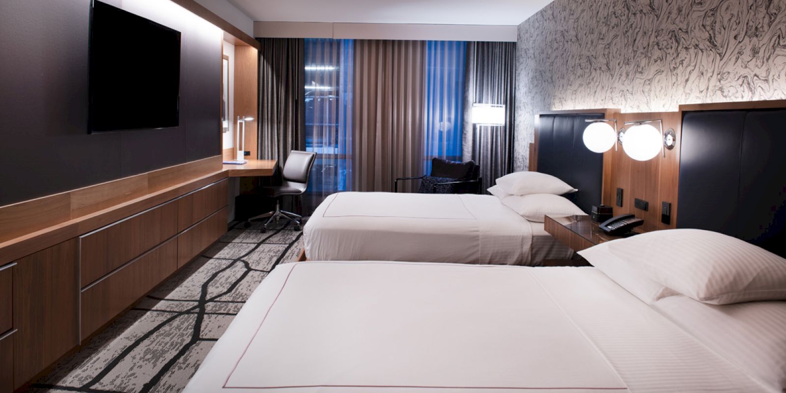 The image shows a modern hotel room with two beds, a flat-screen TV, a desk, and stylish lighting and decor.