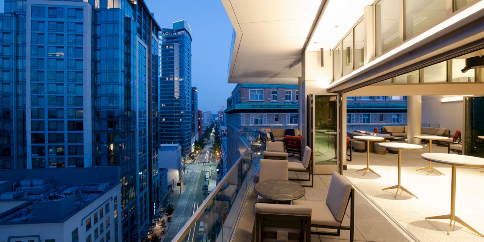 A modern cityscape view from a high-rise balcony with a sleek seating area overlooking the street and neighboring buildings at dusk.