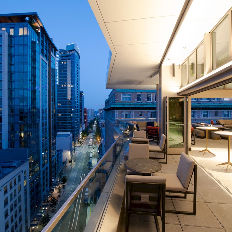 A modern rooftop terrace with seating overlooks a city street and skyscrapers at dusk.