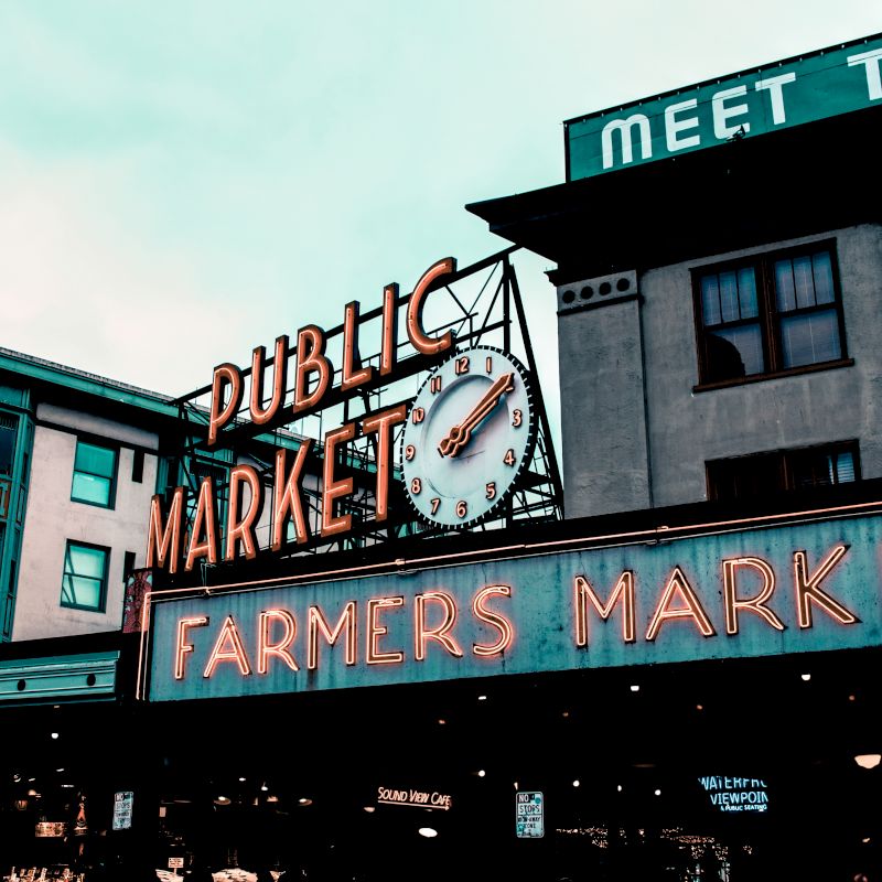 The image shows a building with neon signs reading "Public Market" and "Farmers Market," featuring a large clock above the entrance.