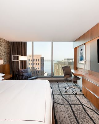 A modern hotel room with a large bed, desk, chair, TV, and window view of city buildings and water, complete with stylish decor.