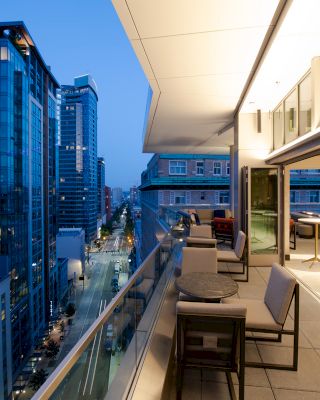 A modern cityscape view featuring a balcony with seating, overlooking a street and several tall buildings at dusk.
