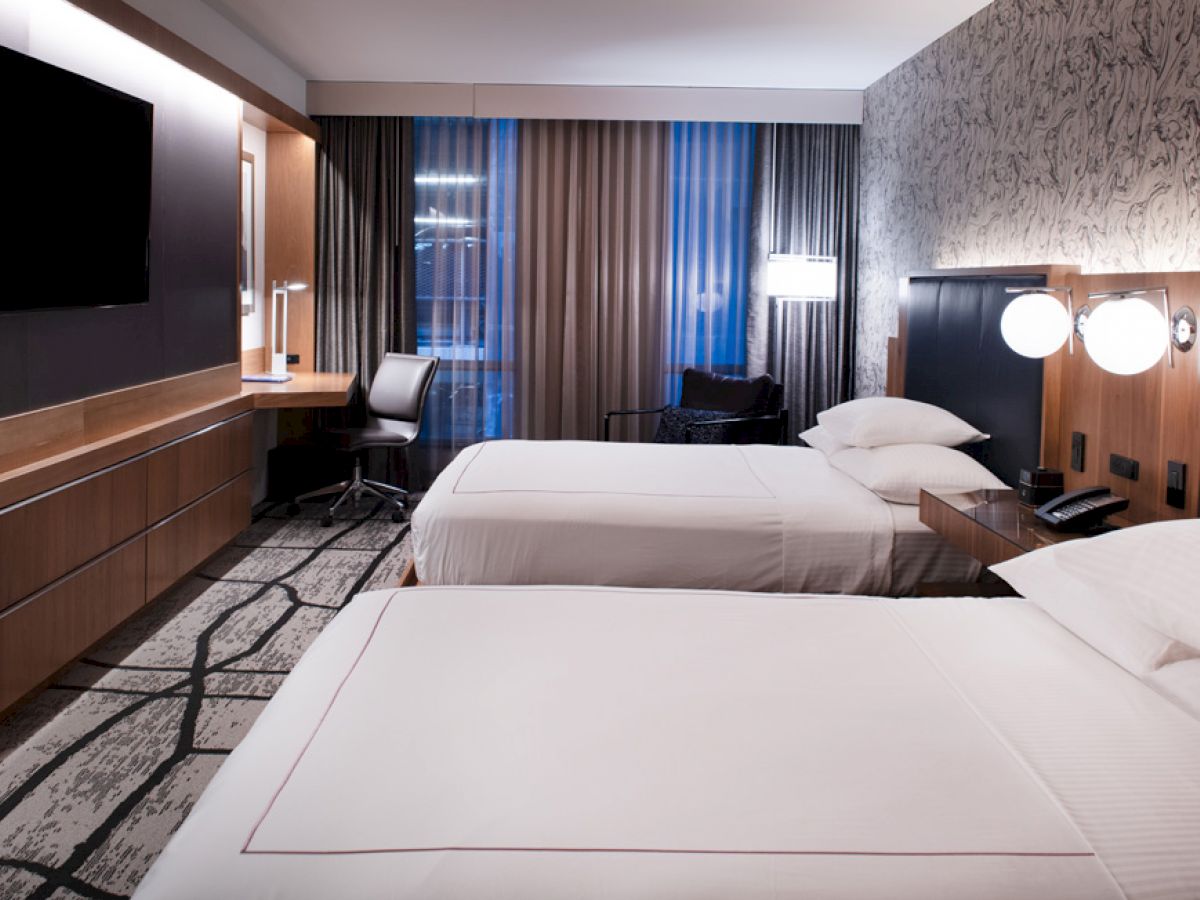 The image shows a modern hotel room with two beds, a wall-mounted TV, a desk, and large windows with curtains.