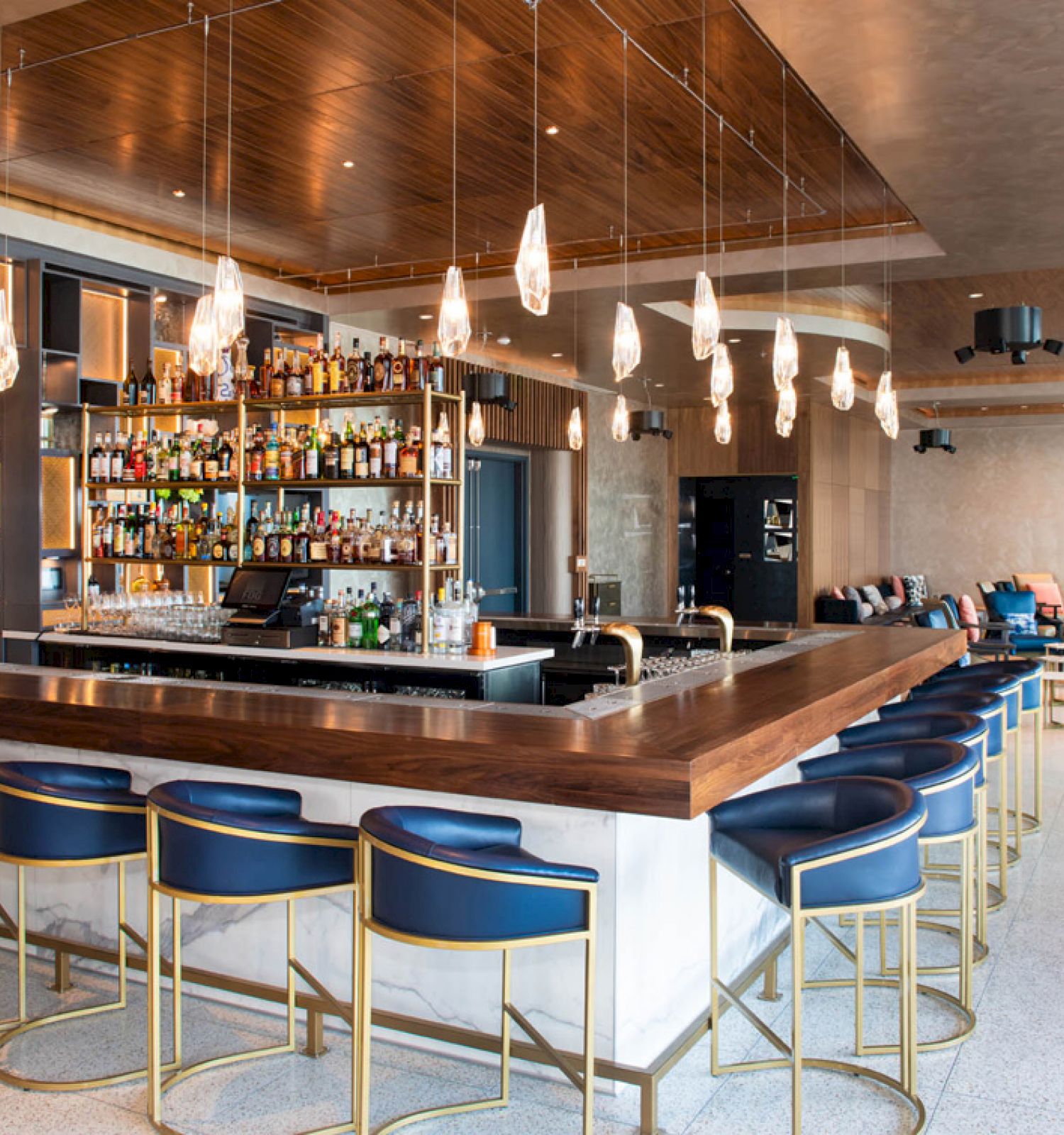 A modern bar with a central counter, surrounded by blue chairs, features a well-stocked back shelf and stylish hanging lights.