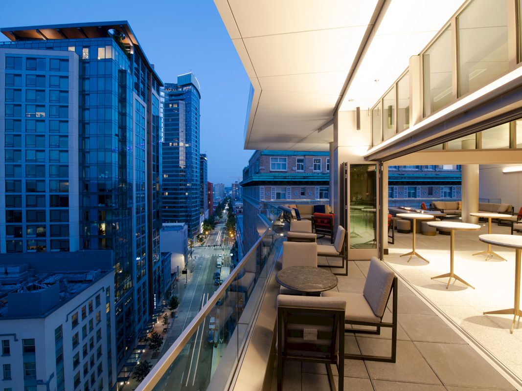 A modern balcony overlooks an urban street with tall buildings at dusk, featuring tables, chairs, and warm indoor lighting.