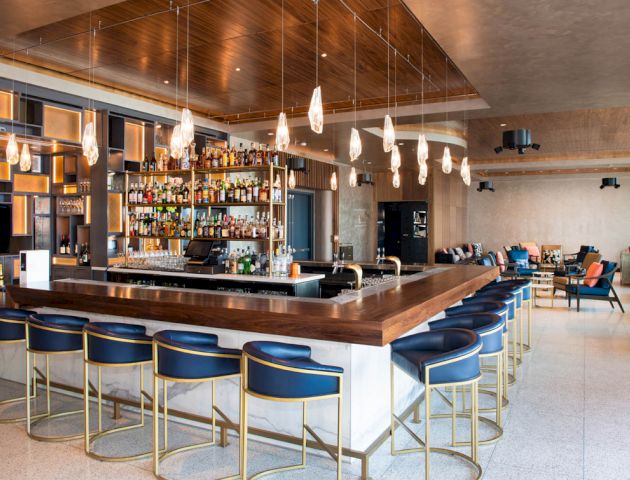 A modern bar with a wooden counter, blue stools, hanging lights, and a well-stocked back shelf in a lounge setting.