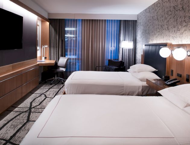 The image shows a hotel room with two beds, a TV, a desk, and modern decor.