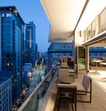 A modern balcony at dusk with seating and view of a cityscape and tall buildings, featuring glass railings and sleek design.
