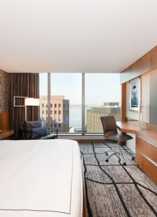 A modern hotel room features a large bed, desk, chair, flat-screen TV, and a window with a city view.