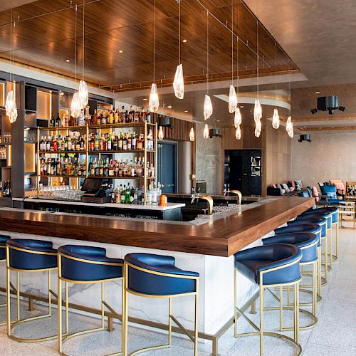 A stylish bar area with hanging lights, blue stools, and a well-stocked drink selection, set in a modern lounge environment.