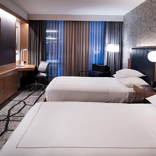 The image shows a modern hotel room with two neatly made beds, a desk with a chair, a wall-mounted TV, lamps, and patterned walls and carpet.