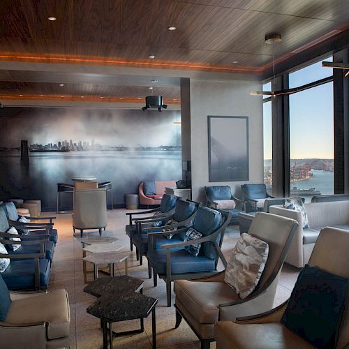 A modern lounge with plush seating and a stunning water view through large windows. A mural of a city skyline decorates one wall.