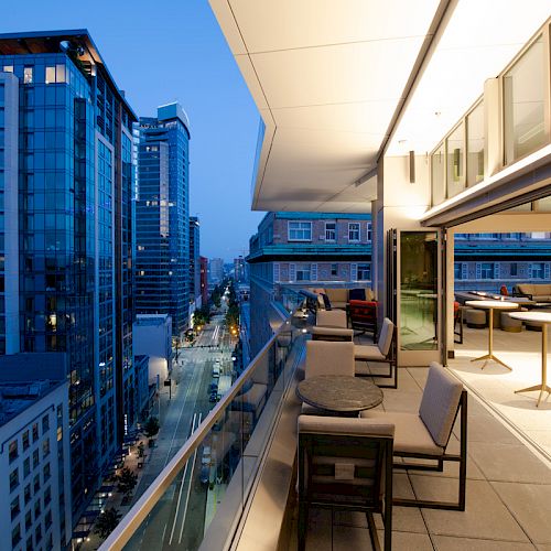 A modern rooftop terrace with seating overlooks a cityscape of tall buildings during twilight, with interior lights on in the adjacent room.