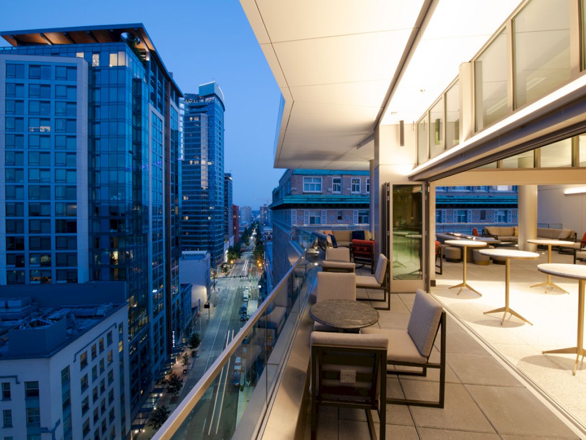 The image shows a modern balcony overlooking a cityscape with tall buildings, featuring seating and a view of the evening skyline.