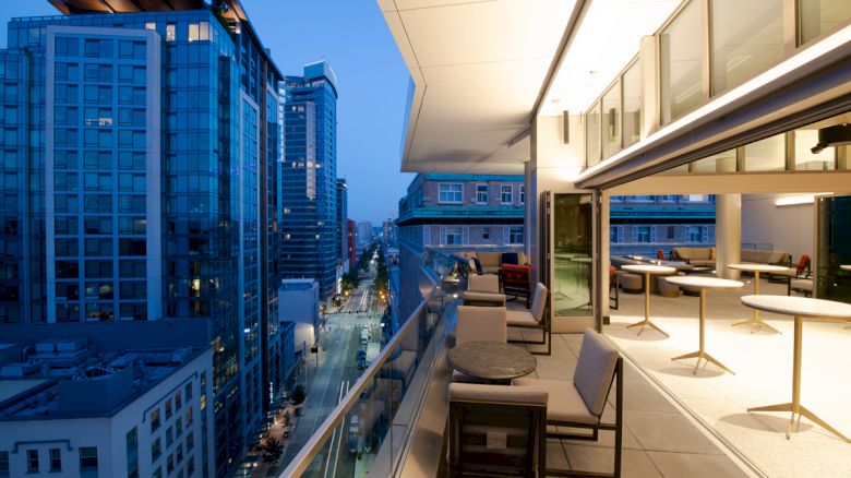 The image shows a modern balcony overlooking a cityscape with tall buildings, featuring seating and a view of the evening skyline.