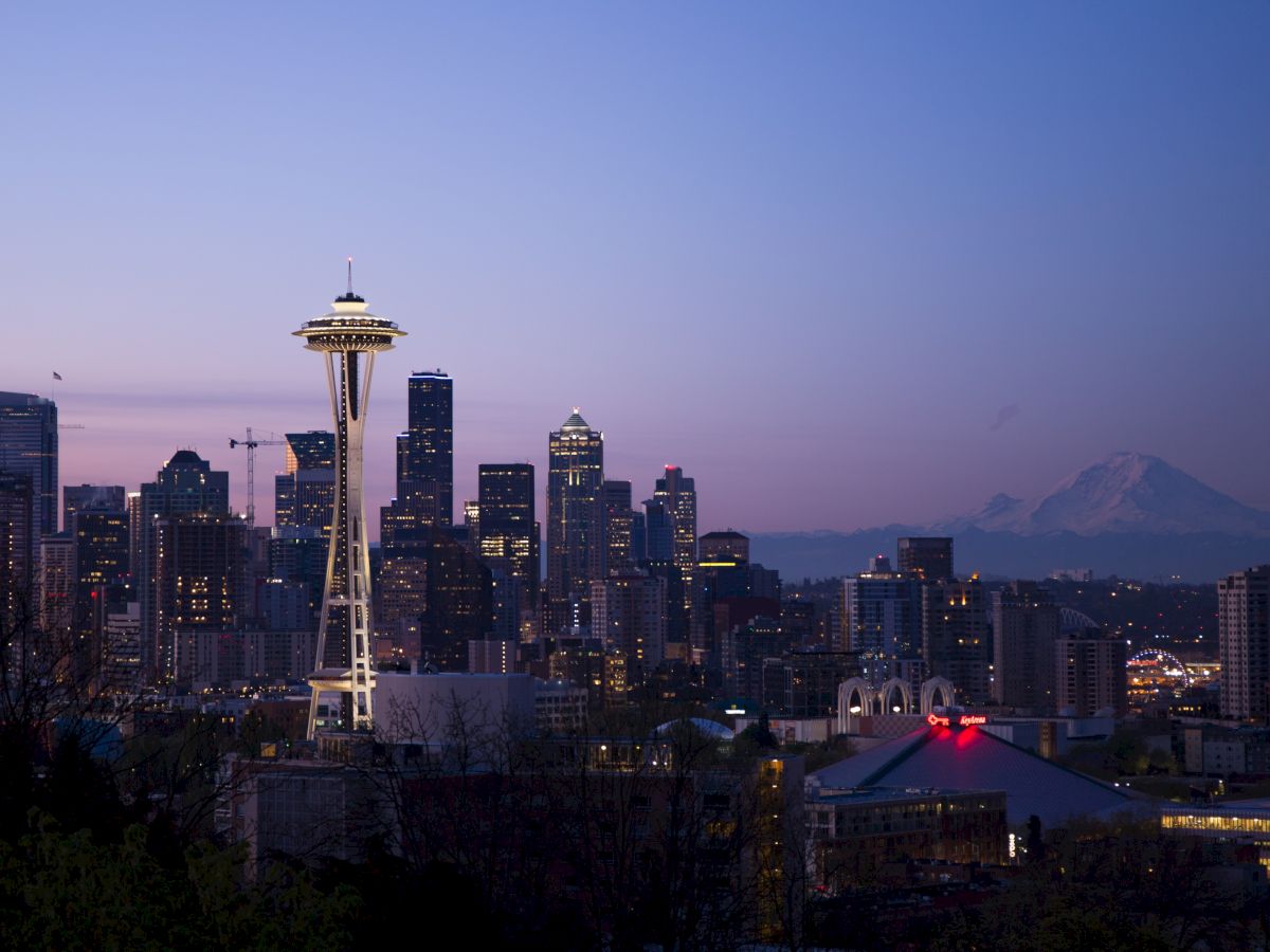 A city skyline at dusk features a tower and surrounding buildings, with a mountain in the distance against a purple-blue sky.