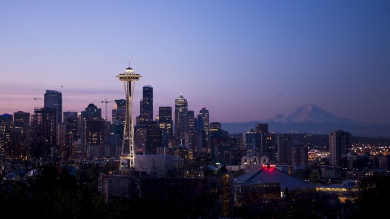 A city skyline at dusk features a tower and surrounding buildings, with a mountain in the distance against a purple-blue sky.
