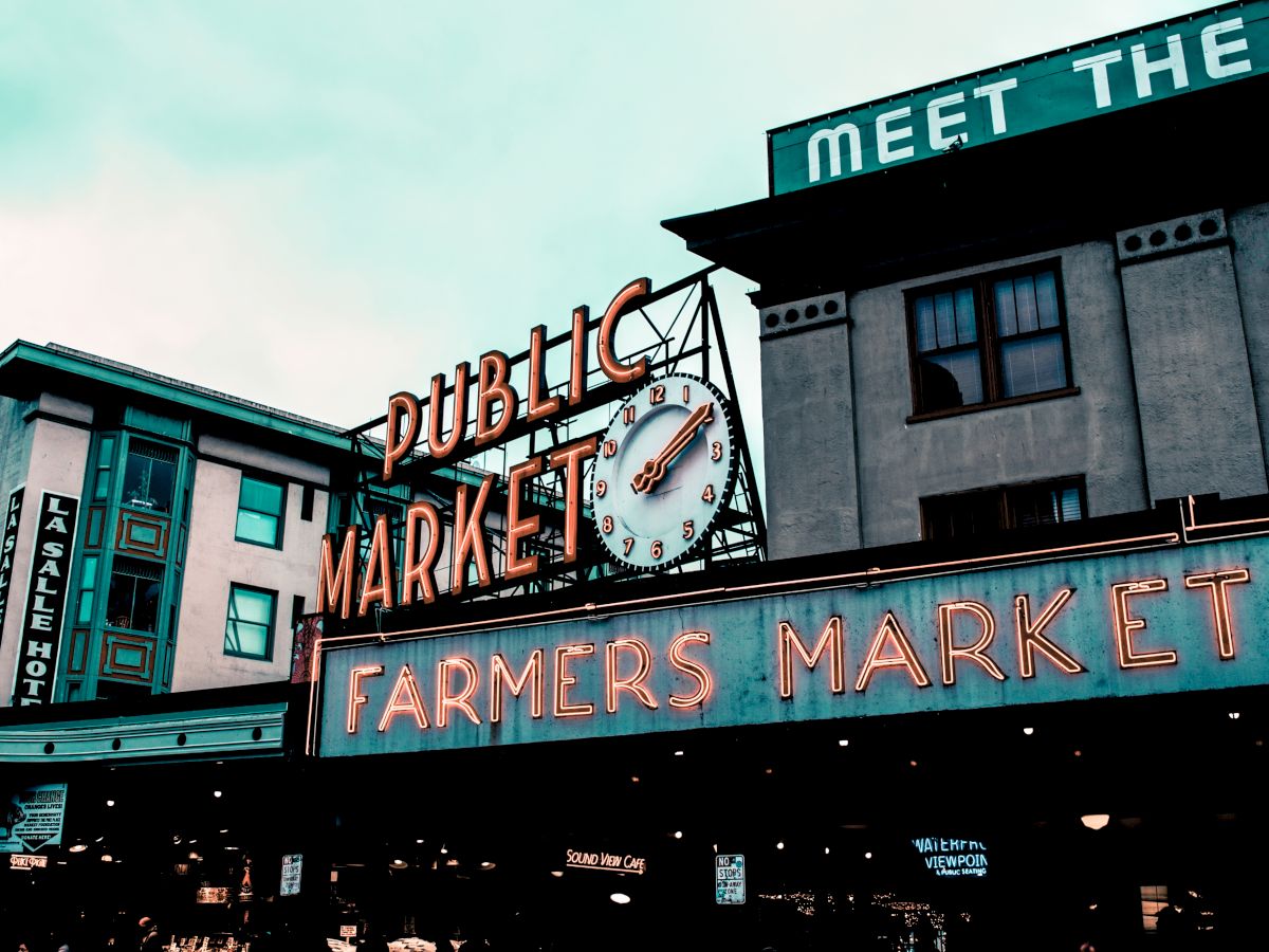 The image shows a neon sign for "Public Market" and "Farmers Market" with a clock, set against urban buildings.