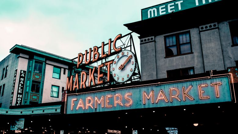 The image shows a neon sign for "Public Market" and "Farmers Market" with a clock, set against urban buildings.