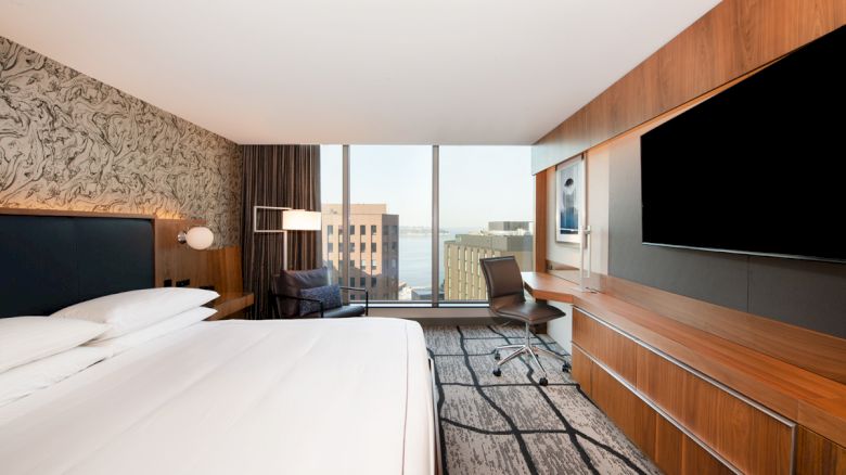 A modern hotel room with a large bed, desk, chair, wall-mounted TV, and city view from the window is shown.