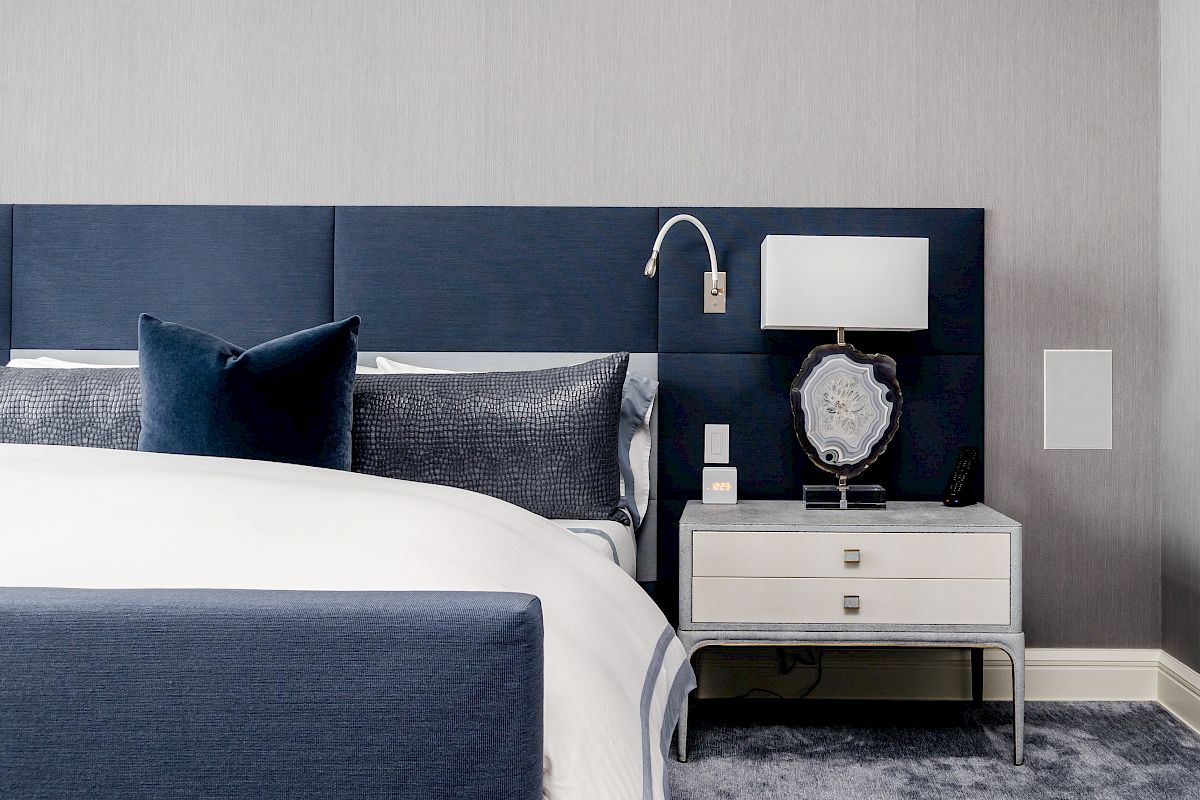 A modern bedroom features a blue headboard, white bedding, and a nightstand with a lamp and decorative item, set on a plush carpet.