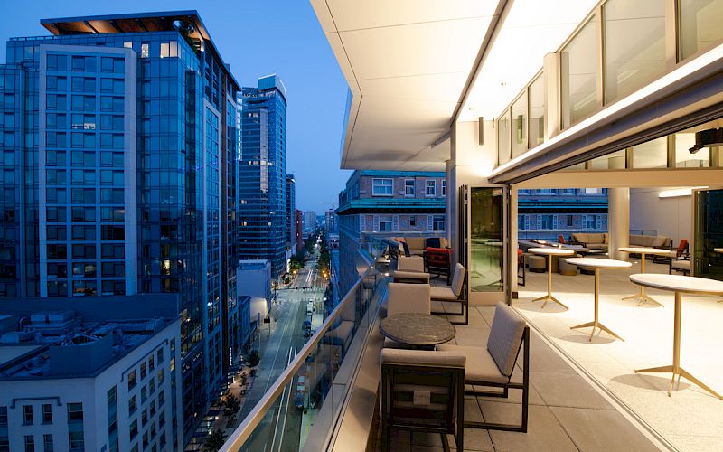 A modern rooftop terrace overlooks a cityscape with tall buildings, featuring outdoor seating and glass railings, lit by soft evening lights.