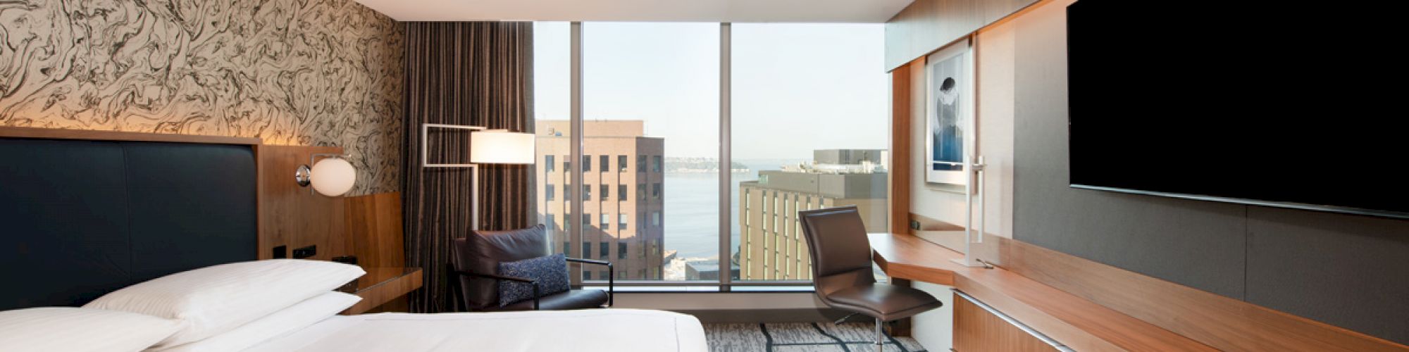 A modern hotel room features a bed, large TV, desk, and a window with a city view. The decor is sleek and contemporary.