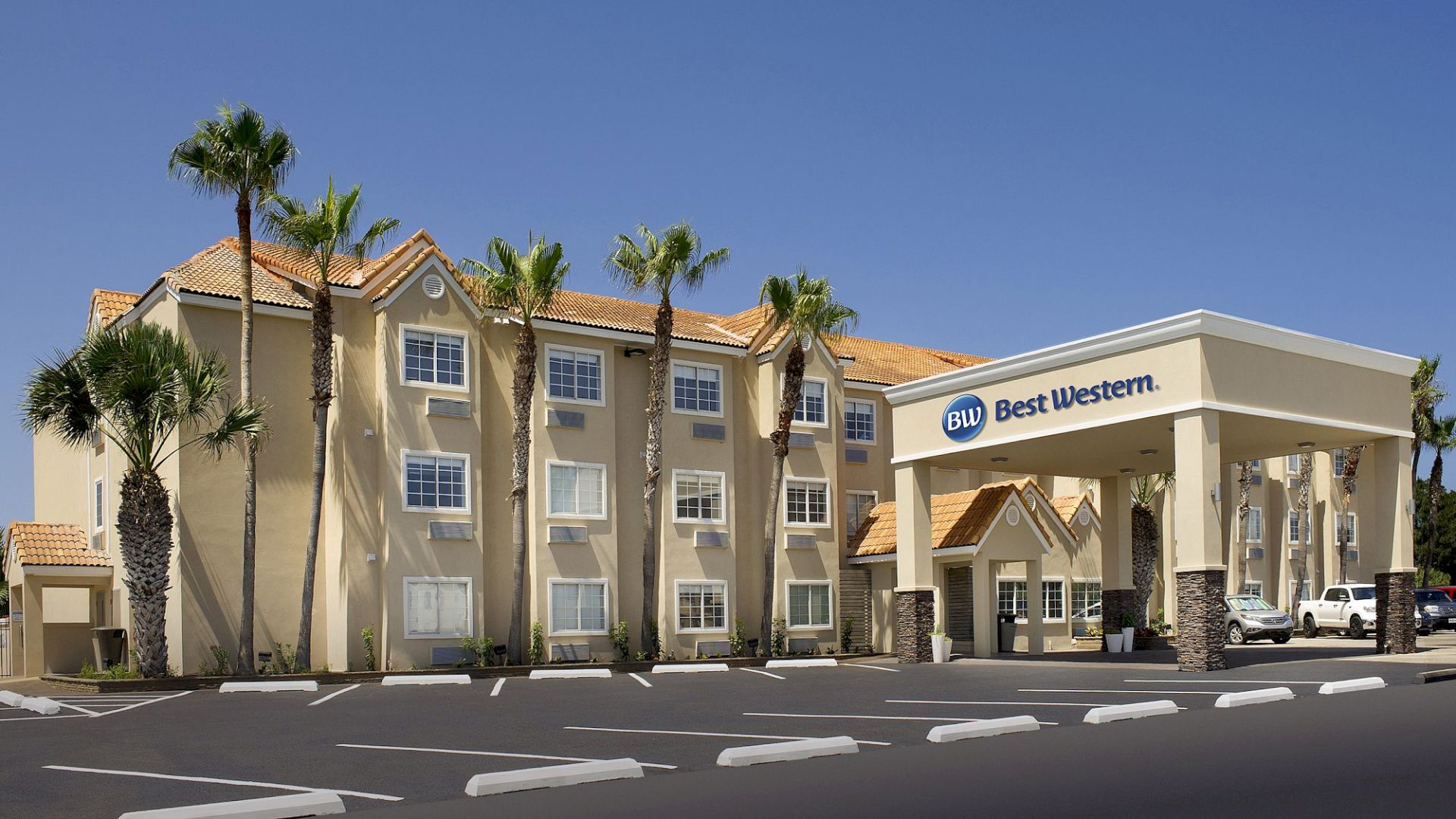 The image shows a Best Western hotel with a beige exterior, palm trees, and a covered entrance area, under a clear blue sky.